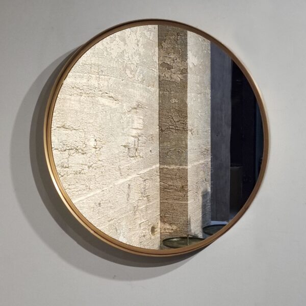De Antique Brass Mirror 87, met een rond ontwerp en een goudkleurig frame, weerspiegelt een zuil van steen of beton met textuur en een deel van een schaduwrijk interieur. Hij is gemonteerd op een effen, lichtgekleurde muur.