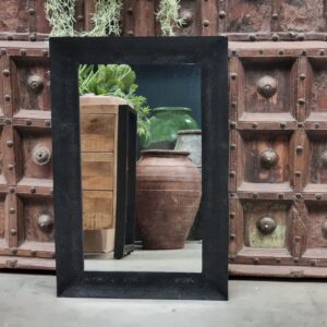 De Spiegel Vierkant Zwart, een grote zwart omlijste spiegel, weerspiegelt weelderige groene planten en hoge keramische vazen die voor een sierlijke rustieke houten deur staan met gedetailleerd geometrisch houtsnijwerk.