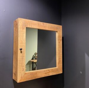 De Badkamerkast Benoa met Deur en Spiegel 80, met een houten frame en een kleine zwarte knop, is gemonteerd op een donkere muur. De spiegel weerspiegelt een deel van een tafel en een lichtgekleurde muur op de achtergrond.