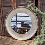 De Spiegel Rond Goud Medium, een ronde spiegel met een brede goudkleurige lijst, rust op een rustiek houten oppervlak en weerspiegelt een modern interieur met grote ramen, stijlvolle meubels en planten die langs één kant groeien.