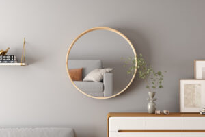 De 'Ria' 80x80 oude eik spiegel weerspiegelt een bank met kussens; onder een dressoir staat een vaas met groene takken, ingelijste kunst en boeken op een zwevende plank tegen een lichtgrijze muur.