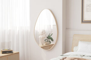 In een knusse slaapkamerhoek hangt de 'Romi' 80x50 Oude eik wandspiegel met een wit borstbeeld en een potplant, bij een raam met transparante gordijnen, een draaiplateau en een keurig opgemaakt bed.