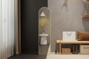 Een moderne minimalistische entree heeft de hoge Spiegel 'Rosalie' 160x40 Oude eik (1c) boogspiegel, een witte kruk met groene takken in een vaas, een geweven bank, neutrale muurkunst, bruine kussens en een Gucci boodschappentas op de vloer.