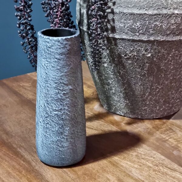 Een Vaasje Langwerpig Blauw (set van 4) vaas staat op een houten oppervlak naast een grotere pot met textuur en donkere bessen en bladeren tegen een blauwgrijze achtergrond.