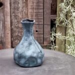 Een set van 4 Vaasje Blauw Gemeleerd, met blauw gestructureerd keramiek met een onregelmatige vorm, staat op een grijs oppervlak tegen rustieke houten planken en hangend groen.