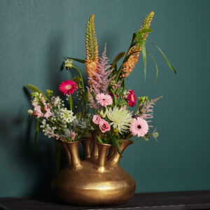 De Vaas Seven Way Antiek goud toont roze gerbera's, witte en gele bloemen en lange oranje aren in zijn drie halzen, op een donker oppervlak tegen een groenblauwe muur.