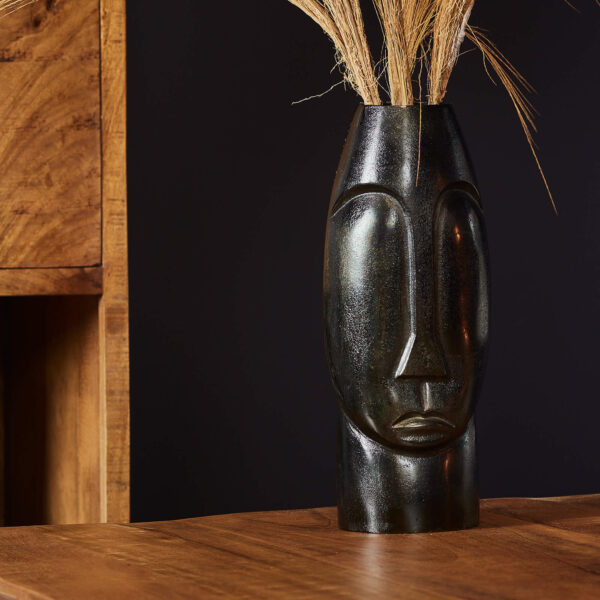 De Vaas Lyra - Zwart | Large, een zwarte vaas met een minimalistisch ontwerp van een menselijk gezicht, houdt gedroogd gras vast op een houten tafel, aangevuld met houten meubels en een donkere muur op de achtergrond.