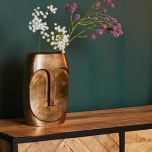 De Vaas Grace - Antiek goud | Large, met een abstract gezichtsontwerp en gevuld met witte en paarse kunstbloemen, staat op een houten kast tegen een donkergroene muur.