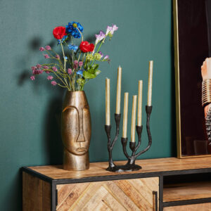 De Vaas Lyra - Antiek goud | Groot, gevuld met kleurrijke bloemen, en een zwarte kandelaar met vijf gouden kaarsen staan op een houten kast tegen een donkergroene muur; een deel van een ingelijste foto verschijnt rechts.