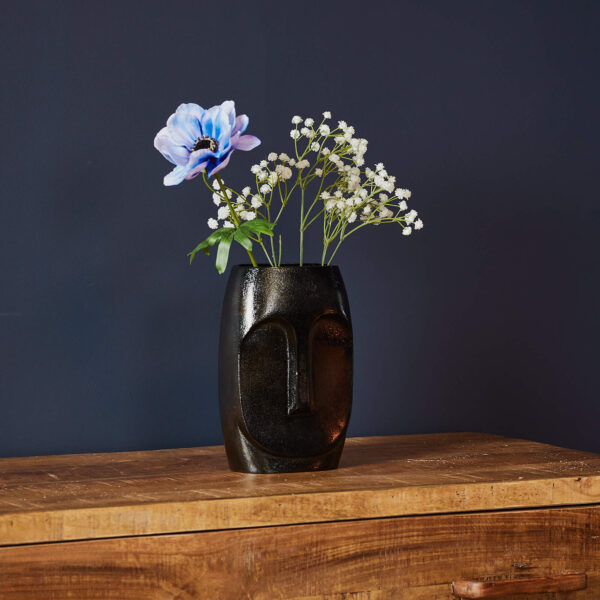 De Vaas Grace - Zwart | Small, een vaas van zwart keramiek met een minimalistisch gezichtsontwerp, houdt een blauwe bloem en witte bloesems op een houten oppervlak tegen een donkerblauwe muur.