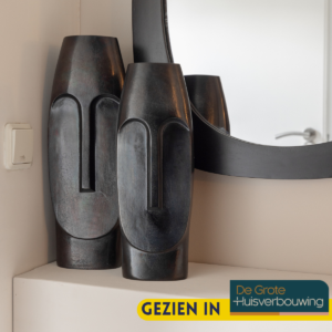 Twee Vaas Liv - Zwart | Grote vazen met abstracte gezichtsontwerpen staan op een witte hoekplank bij een spiegel. Aan de muur hangt een lichtschakelaar en in de hoek prijkt een label Gezien in De Grote Huisverbouwing.