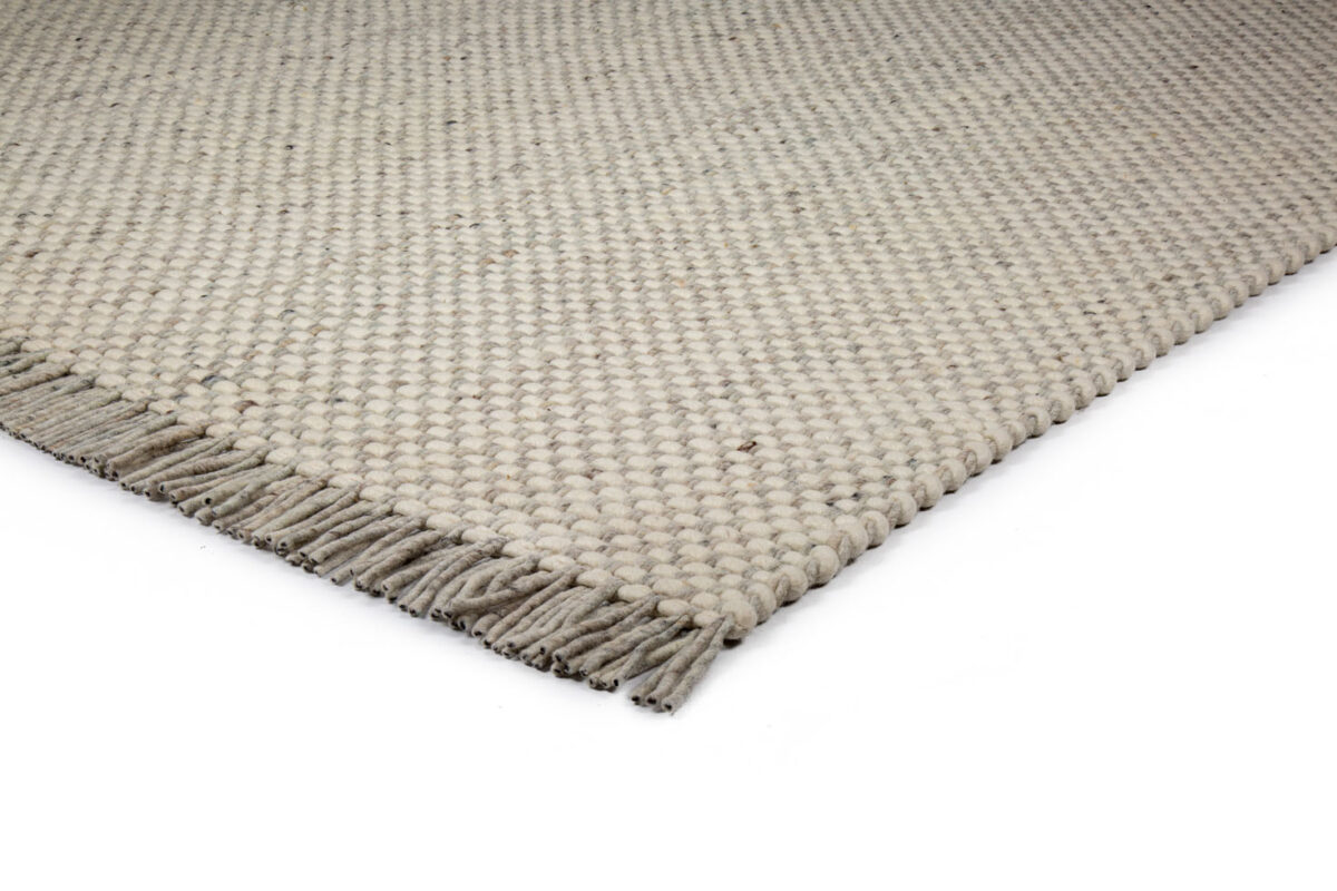 Een close-up van de hoek van het Burano White Grey 001-367 170x230 tapijt, met een lichtbeige grof geweven textuur en korte franjes langs de rand, op een wit oppervlak.