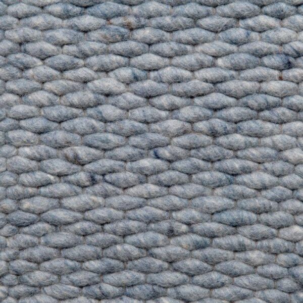 Een close-up van het Genua Sky Blue 227 200x300 tapijt toont de grijze, geweven wollen textuur met dikke, vervlochten garens in een zacht, licht pluizig horizontaal patroon.