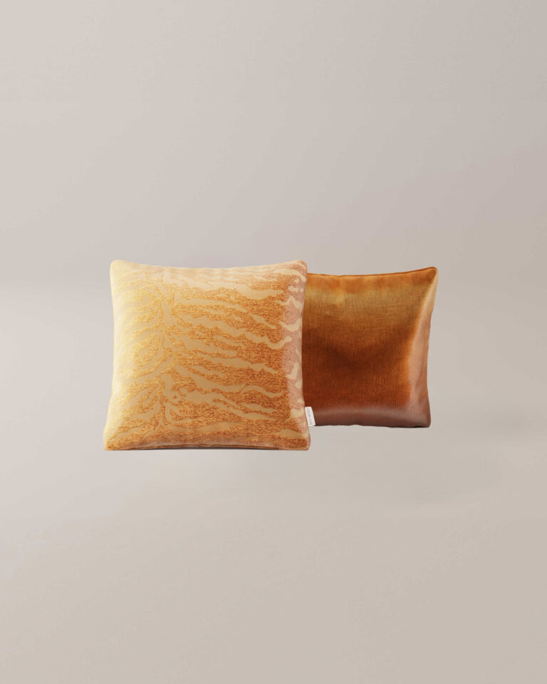 Twee vierkante Cognac hoofdkussens - de ene met een gestructureerd geeloranje patroon en de andere in gladde, glanzende koperbruine stof - liggen naast elkaar tegen een neutrale achtergrond.