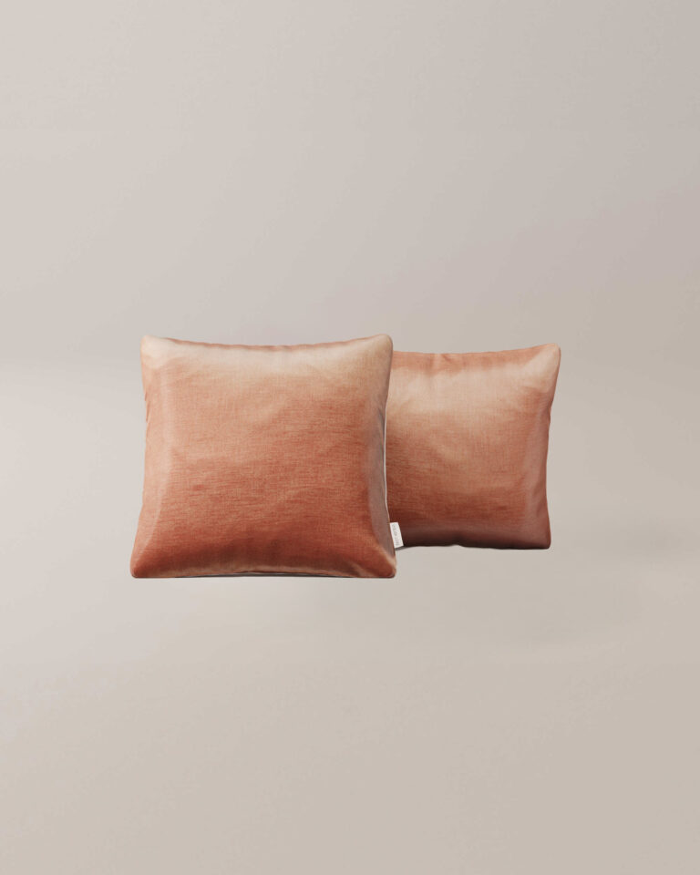 Twee vierkante sierkussens "Rose" met een zacht kleurverloop van perzik naar roest tegen een effen lichtbeige achtergrond.