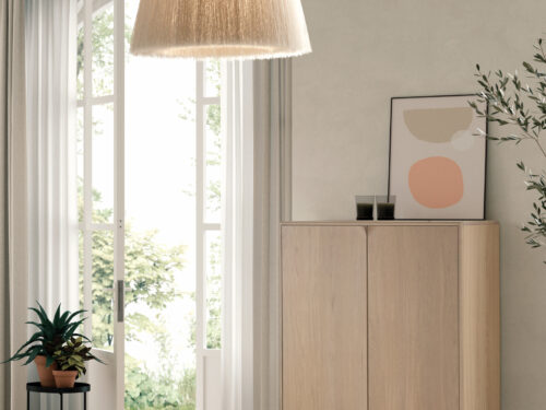 Een moderne, minimale woonkamer met de SENTO BERGKAST 2 WWM lichte houten kast, twee hanglampen, abstracte kunst, twee glazen op de kast, een plant in pot op een zwarte standaard en grote open ramen met transparante witte gordijnen.
