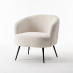 De Laura fauteuil Stof Teddy Ivory 101 is een moderne fauteuil met een gebogen rugleuning, bekleed met ivoorkleurige Teddy-stof en voorzien van vier slanke, schuine zwarte poten tegen een effen witte achtergrond.