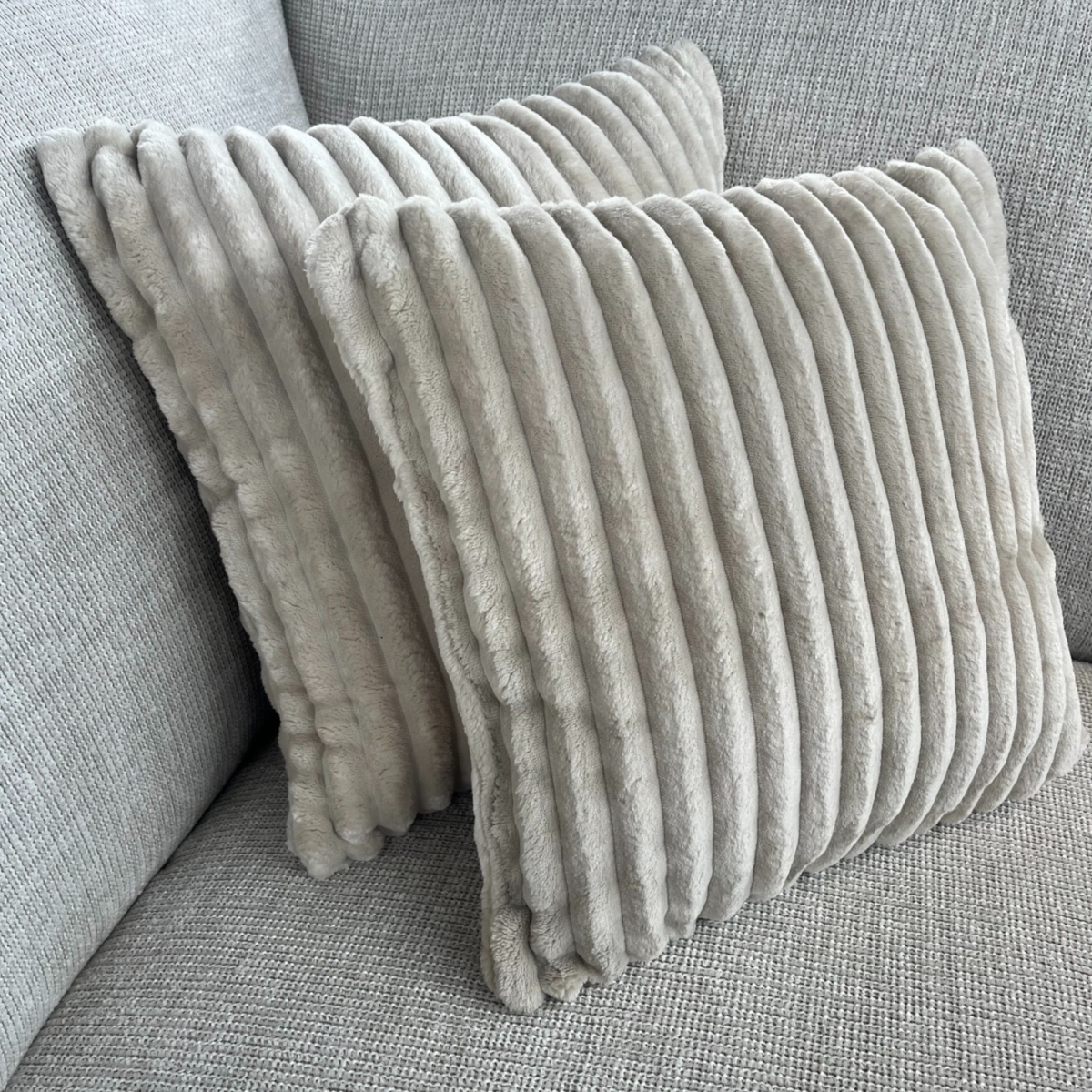 Twee MySofa Kussens | Tilia 03 | 40x40 cm in lichtgrijs, met een geribbeld pluche design, liggen op een bijpassende lichtgrijs gestoffeerde bank, schuin geplaatst met het ene kussen iets voor het andere.