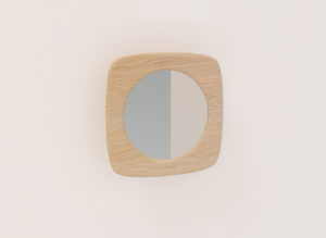 De SENTO SPIEGEL90 E02M, een ronde spiegel met een licht houten vierkant frame, is gemonteerd op een effen, lichtgekleurde muur en weerspiegelt een subtiele verticale verdeling van licht en schaduw.