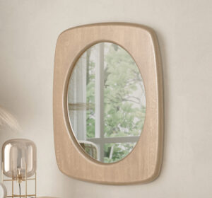 De SENTO SPIEGEL90 WWM, een ovale spiegel met houten lijst, hangt aan een lichte muur en weerspiegelt een raam met groen. Een decoratieve lamp en verschillende voorwerpen zijn gedeeltelijk zichtbaar in de linkerbenedenhoek.