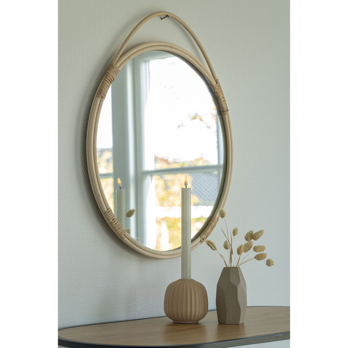 De Malo Mirror, een ronde spiegel van natuurlijk rotan (Ø50 cm), hangt aan een witte muur boven een houten tafel met een brandende witte kaars in een geribbelde houder en een vaas met gedroogd gras. Een raam wordt weerspiegeld in de spiegel.