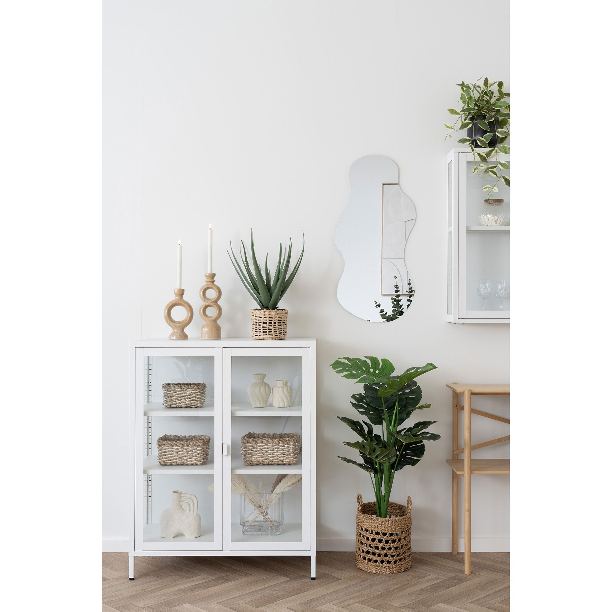 Een moderne witte kast met glazen deuren bevat gevlochten manden en decor, met daarboven kandelaars en een potplant. Vlakbij staat een grote monstera. De minimalistische kamer is afgewerkt met een wandplank, een stoel en de Palermo spiegel.