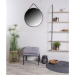 Minimalistische kamer met de Trapani spiegel (zwart stalen frame, PU band, Ø60 cm), potplant in een rieten mand, grijs kussen op een bank, houten planken met decor, zwarte lantaarn, decoratieve schaal en rond grijs vloerkleed op lichthouten vloer.