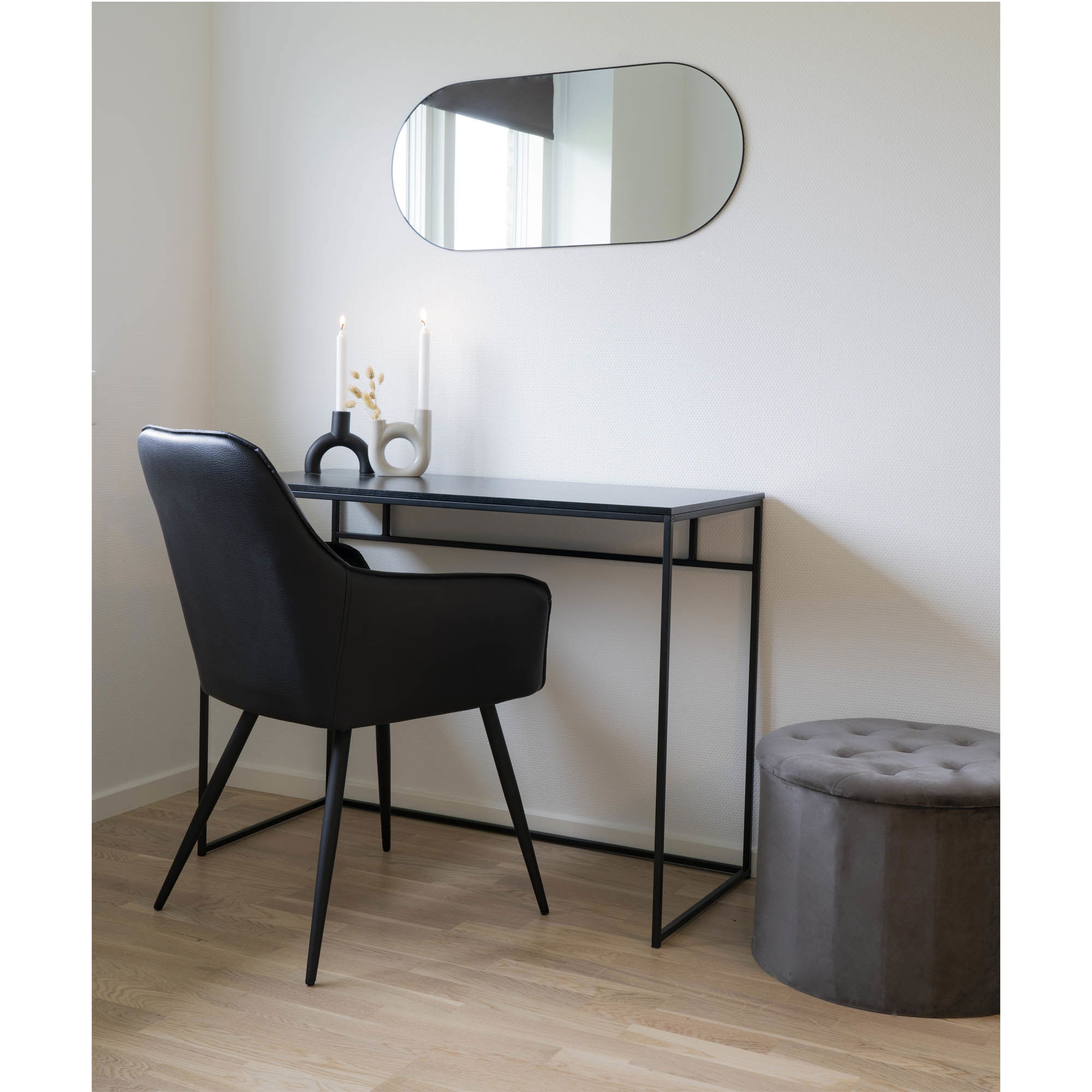 Een minimalistische werkplek heeft een zwarte stoel aan een slank zwart bureau met twee brandende kaarsen en decor, een getufte grijze voetenbank vlakbij en de Jersey Mirror Oval - een ovale spiegel met een zwarte lijst van 35x80 cm - aan de muur erboven.