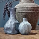 Twee Vaasje Blauw Gemeleerd vazen uit een set van 4, met blauw getextureerd glazuur, staan op een houten ondergrond voor een grote, rustieke pot. Een tak met donkere bessen steekt uit de pot op de achtergrond.