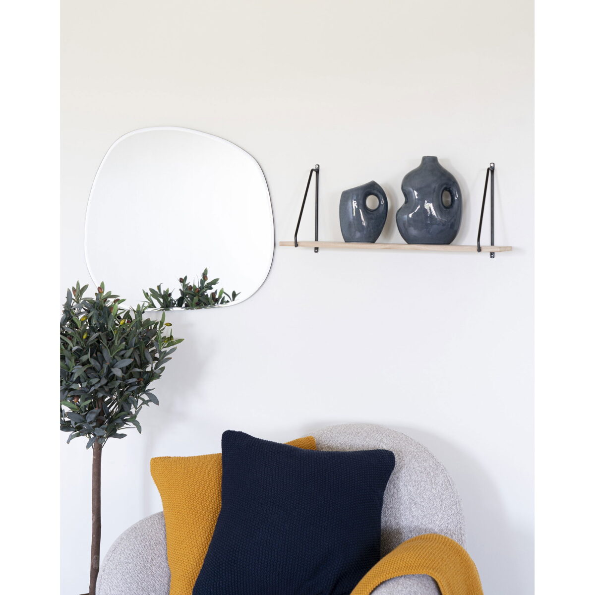 Een gezellig hoekje met een lichtgrijze stoel, kussens in navy en mosterd, een bladplant, een onregelmatige wandspiegel en een houten plank met twee moderne blauwe vazen.