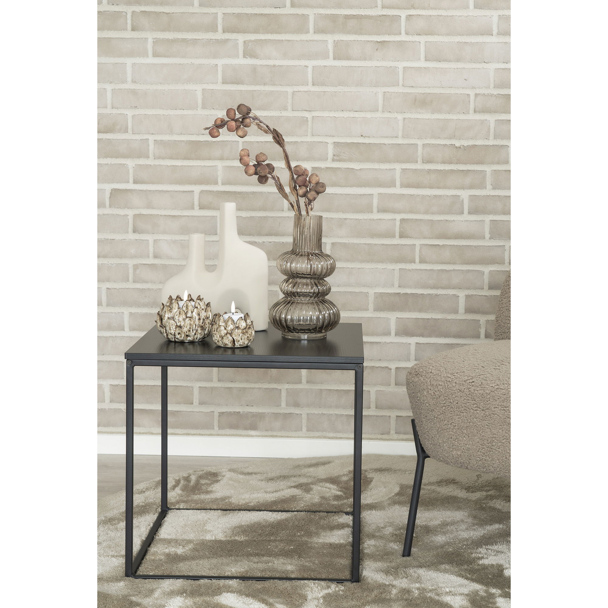 Op een zwart metalen bijzettafeltje staan twee metalen kaarsenhouders en de vaas van gerookt mondgeblazen glas Ø15x25 cm met gedroogde takken, plus een vaas van wit keramiek, naast een stoel met beige textuur tegen een lichte bakstenen muur.