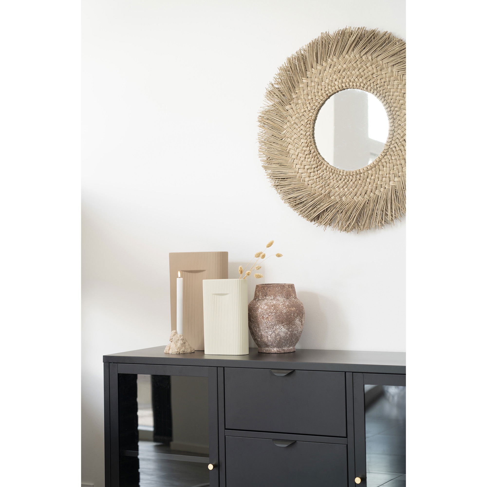 Een zwarte kast met gouden knoppen is bedekt met een beige kandelaar, een geribbelde vaas, een vaas van grijs keramiek (16,5x6,5x26 cm) en gedroogde bloemen, geplaatst onder een ronde geweven spiegel op een witte muur.