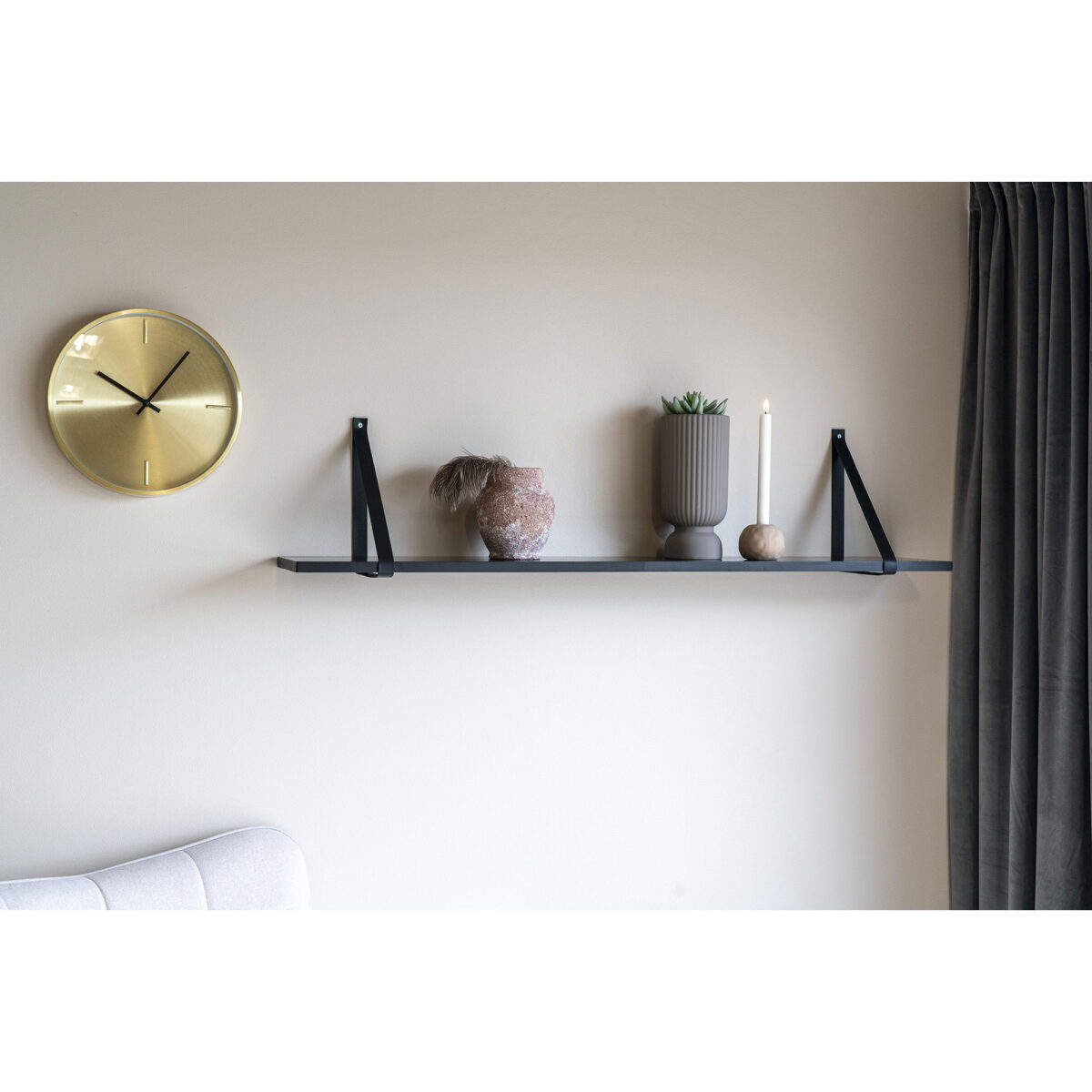 Een gouden wandklok, een zwevende zwarte plank met een vaas van grijs keramiek (Ø12x25,5 cm), een grijze potplant, een kaars en een klein rond object staan op een beige muur met een donker gordijn aan de rechterkant.