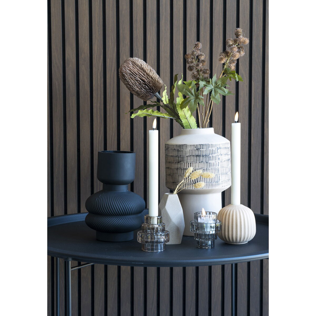 Een modern tafelblad met een witte vaas met gedroogde bloemen, twee brandende witte kaarsen in glazen houders, een kleine geribbelde beige kaars, een vaas van zwart glas Ø15x22 cm en een geometrisch ornament tegen verticale houten panelen.