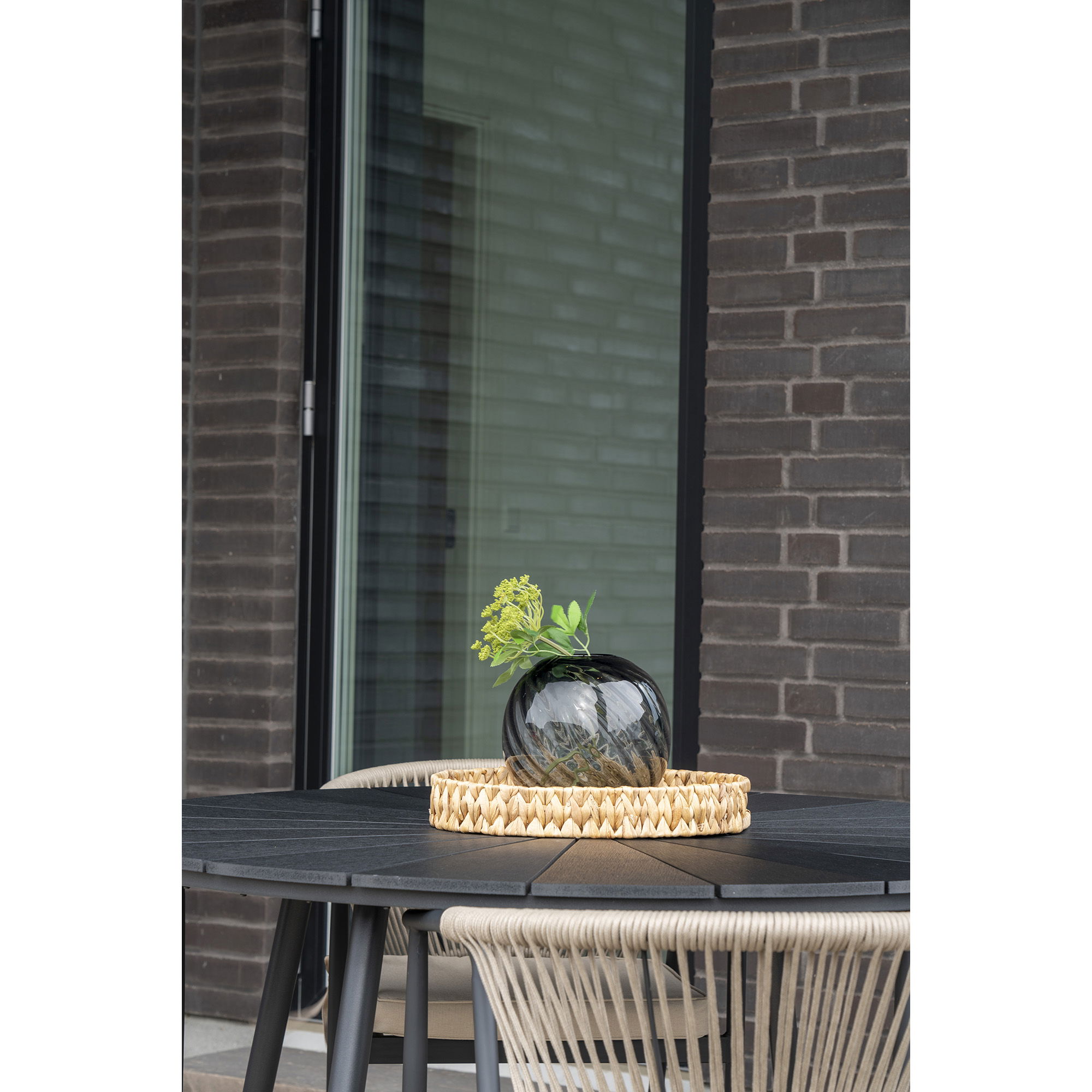Een zwarte ronde tafel met touwgeweven stoelen staat bij een bakstenen muur. Daarop staat een geweven dienblad met een mondgeblazen vaas van grijs glas (Ø20x19 cm) met groen gebladerte. Een glazen deur en raam verschijnen op de achtergrond.