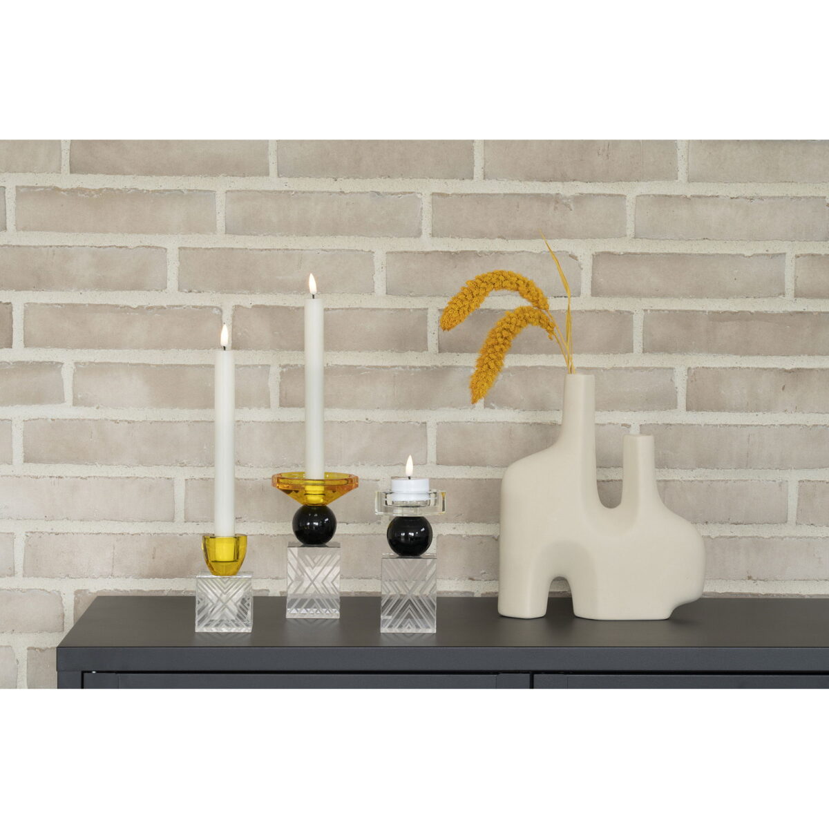 Een minimalistische display met twee brandende kaarsen in moderne houders en een vaas van zandkeramiek met twee openingen (8x23,5x27 cm) met gedroogde tarwe, alles op een donker oppervlak tegen een lichte bakstenen muur.