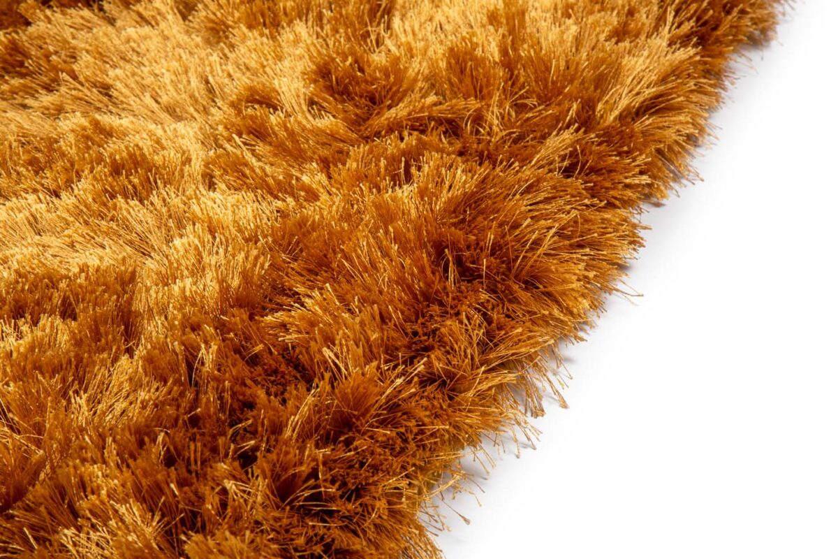 Close-up van het Arezzo ST Gold 048 170x230 tapijt met goudbruine, lange, dikke vezels tegen een witte achtergrond. De zachte, pluche textuur en dichte, pluizige pool worden benadrukt voor een luxueuze uitstraling.