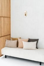 Een minimalistische sofa met vijf Sierkussenhoes Scotty Donkerbruin 50x50 cm kussens staat tegen een witte muur met een moderne wandlamp erboven. Aan de linkerkant van de afbeelding staat een houten lattenwand.