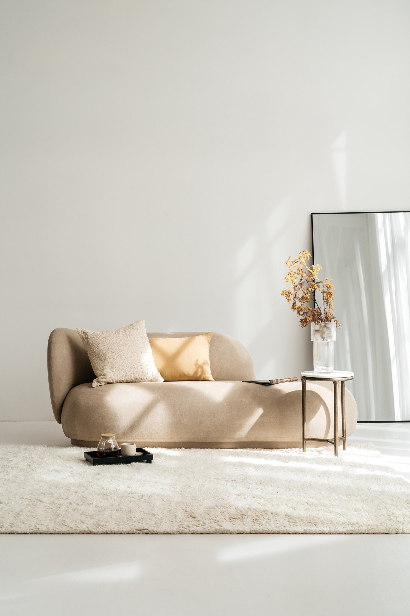 Een minimalistische woonkamer met een beige bank, het Sierkussenhoes Scotty Goudbruin 60x40 cm kussen, een klein bijzettafeltje met droogbloemen in een vaas, een dienblad met een drankje, een ruig wit vloerkleed en een grote spiegel tegen de witte muur.