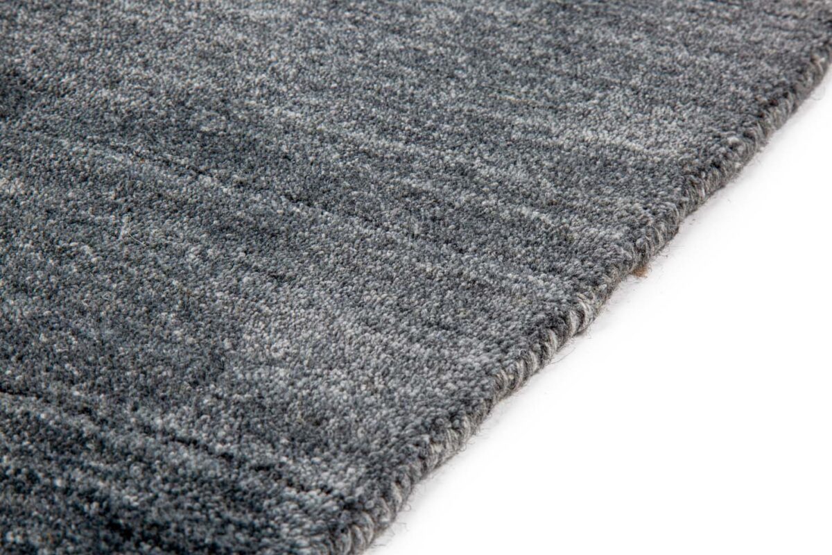 Close-up van de hoek van het Portofino Blue 01 200x300 vloerkleed op een witte achtergrond. Het zachte, geweven oppervlak met subtiele kleurvariaties en een dikke, afgewerkte rand zijn zichtbaar.
