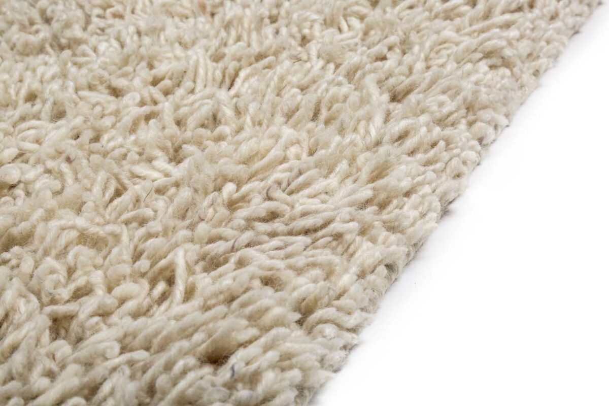 Close-up van de hoek van het Berbero Lungo Cloud White 815 170x230 tapijt op een witte achtergrond, met de nadruk op de dikke beige vezels met textuur.
