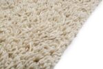 Close-up van de hoek van het Berbero Lungo Cloud White 815 170x230 tapijt op een witte achtergrond, met de nadruk op de dikke beige vezels met textuur.