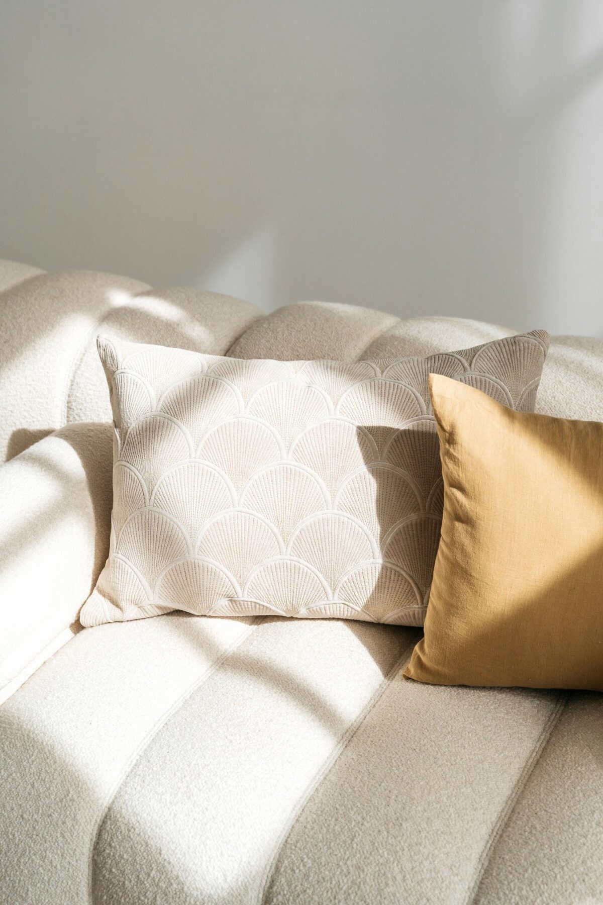 Een crèmekleurige bank met twee kussens: de ene heeft een subtiel schulppatroon in lichtbeige en de andere is het Sierkussenhoes Scotty Goudbruin 50x50 cm in goudbruin, beide verlicht door zacht natuurlijk licht.