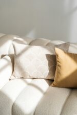 Een crèmekleurige bank met twee kussens: de ene heeft een subtiel schulppatroon in lichtbeige en de andere is het Sierkussenhoes Scotty Goudbruin 50x50 cm in goudbruin, beide verlicht door zacht natuurlijk licht.