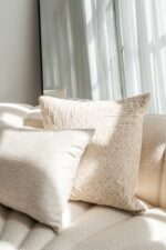 Twee Sierkussenhoes Lynn 50x50 cm met beige structuur liggen op een crèmekleurige bank, met binnenvallend zonlicht en een grote spiegel op de achtergrond die het licht weerkaatst.