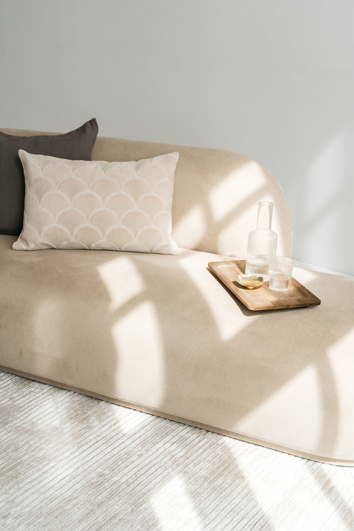 Op een beige bank staat het Sierkussenhoes Jane 60x40 cm, gecombineerd met een houten dienblad met twee glazen, een karaf en schijfjes citroen. Zonlicht werpt zachte schaduwen en creëert een rustige en minimalistische sfeer.