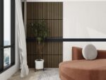 Minimalistische woonkamer met een bruine gebogen bank, rond grijs kussen, plant in pot, vloerkleed met patroon en een Akupanel Eiken Walnoot 60 x 60 cm Lattenwand Akoestische wandpanelen accentmuur. Een groot raam met witte gordijnen brengt natuurlijk licht binnen.