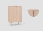 De SENTO BERGKAST 2 WWM is een licht houten kast met twee deuren en vier poten tegen een effen achtergrond. Een close-up inzet toont het greepontwerp, met naar binnen gebogen deurranden voor een elegante, subtiele greep.