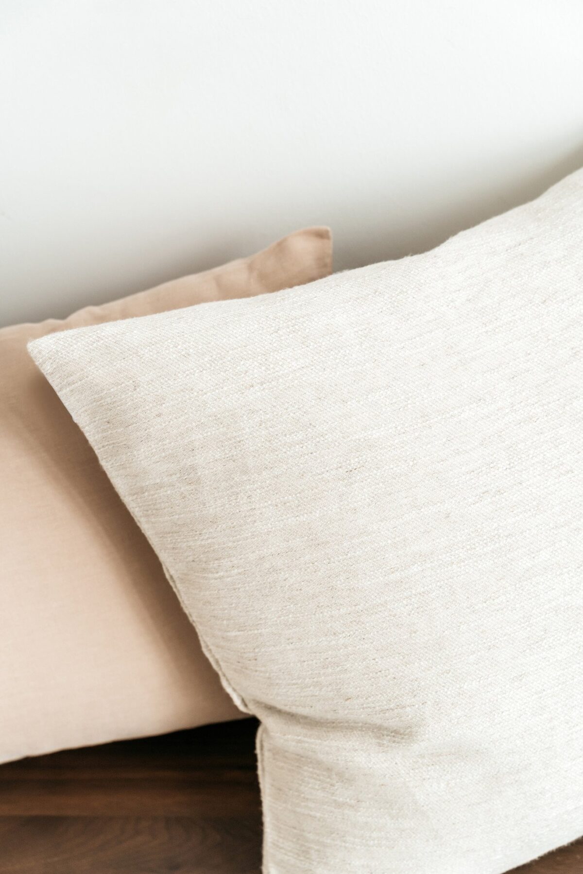 Twee kussens liggen op een bed tegen een witte muur; op de ene ligt de hoes van het Sierkussenhoes Hazel 50x50 cm in lichtbeige, op de andere een zachtbruine hoes. Een donker houten oppervlak is gedeeltelijk zichtbaar.