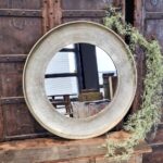 De Spiegel Rond Goud Medium, met zijn metalen frame, hangt aan een rustieke houten muur en weerspiegelt een groot raam en het buitenlandschap. Eén kant van het frame is versierd met gedroogd groen voor extra charme.